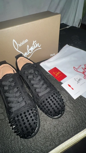 Christian Louboutin svarta sneakers med nitar - Svarta sneakers från Christian Louboutin med coola svarta nitar på tån och klassisk röd sula. Skorna har snörning och är gjorda i mocka med detaljer i skinn. Perfekt för dig som vill sticka ut med en lyxig och edgy stil.