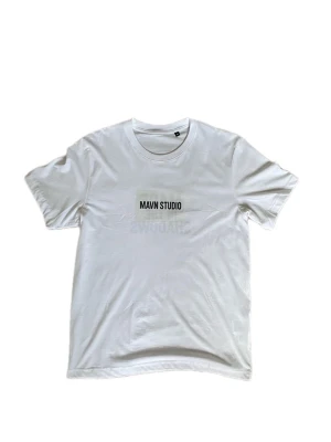 Vit t-shirt från MAVN Studio - Snygg vit t-shirt från MAVN Studio med svart tryck på bröstet och ryggen. Framtill står det 'MAVN STUDIO' och på ryggen 'MADE IN THE SHADOWS'. Klassisk rund hals och korta ärmar. Perfekt för dig som gillar cleana och stilrena plagg med statement-tryck.