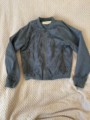 Blå bomberjacka med broderade blommor för barn - Snygg blå bomberjacka för barn från POMP de LUX med broderade blommor på framsidan. Jackan har dragkedja, ribbade muddar och två fickor med dragkedja. Materialet är polyester och jackan är långärmad med en loose passform.