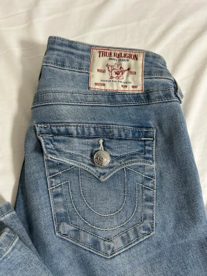 True Religion Joey Low Rise Flare Jeans - Säljer ett par ljusblå True Religion Joey jeans med låg midja och flare-ben. Klassiska kontrastsömmar, stora fickor med lock och silverknappar. Ikonisk True Religion patch bak och snygg tvättad denim-look. Perfekta för dig som gillar retrovibbar och statement-jeans.