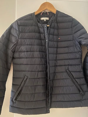 Svart pufferjacka Tommy Hilfiger XS - Svart pufferjacka från Tommy Hilfiger i storlek XS. Jackan har ett stilrent, quiltat mönster, rund hals och två fickor med dragkedja. Liten logga på bröstet och diskreta knappar framtill. Perfekt för kyliga dagar och enkel att matcha med din outfit.
