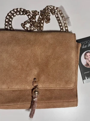 Beige axelväska i mocka med kedja - Säljer en snygg beige axelväska från Amara i mjuk mocka. Väskan har en stilren design med guldfärgad kedjerem och dekorativ tofs framtill. Perfekt storlek för det viktigaste och passar till många outfits.