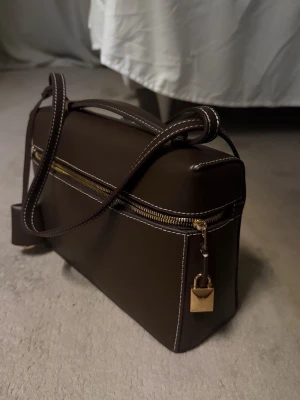 Brun axelväska med gulddetaljer - Snygg brun axelväska i skinn med vit kontrastsöm och guldfärgad dragkedja. Väskan har en boxig form, justerbar axelrem och ett dekorativt hänglås i guld. Perfekt för dig som gillar stilrena accessoarer med lyxig känsla.