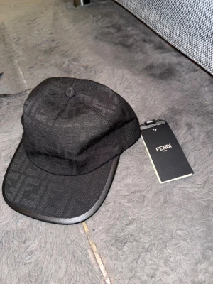 Svart Fendi keps med logomönster - Snygg svart keps från Fendi med diskret logomönster över hela ytan. Justerbar rem baktill med metallspänne och Fendi-logga. Materialet är bomull och polyester, vilket gör den både bekväm och stilren. Perfekt för dig som vill ha en exklusiv accessoar med streetkänsla.
