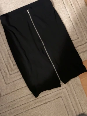 Svart kjol med dragkedja framtill - Snygg svart kjol med en lång, silvrig dragkedja som går hela vägen framtill. Kjolen har en rak och stilren form, perfekt för dig som gillar minimalistisk design. Materialet känns slätt och bekvämt mot huden. Enkel att matcha med olika toppar och accessoarer.