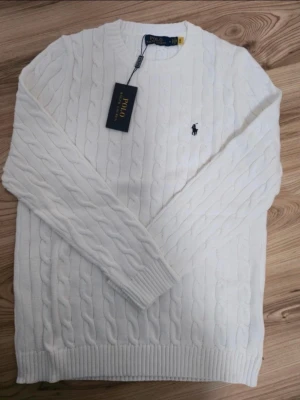 Vit stickad tröja från Polo Ralph Lauren - Säljer en vit stickad tröja från Polo Ralph Lauren med klassiskt kabelstickat mönster och rund hals. Tröjan har långa ärmar och den ikoniska logon broderad på bröstet. Perfekt för dig som gillar stilrena och tidlösa plagg.