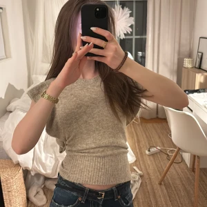 Beige stickad topp med volangärm - Säljer en beige stickad topp med volangdetaljer på axlarna och ribbad nederkant. Toppen är ärmlös och har en rund halsringning. Perfekt att styla med jeans eller kjol för en chill look.