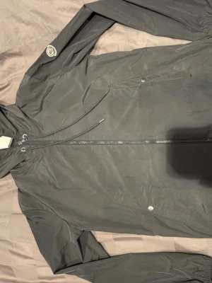 Grå vindjacka från Moncler - Snygg grå vindjacka från Moncler med huva och dragkedja framtill. Jackan har vita, röda och marinblå ränder på huvan samt Moncler-logga på ärmen. Tillverkad i lätt polyester, perfekt för blåsiga dagar. Fickor med knappstängning och justerbar dragsko vid huvan.