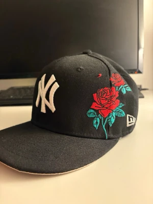 Svart New Era keps med rosbroderi - Säljer en svart New Era keps med New York Yankees-logga i vitt framtill och snyggt broderade röda rosor med gröna blad på sidan. Kepsen har även ett färgglatt World Series-märke på sidan och är tillverkad i bomull. Perfekt för dig som vill sticka ut med coola detaljer.