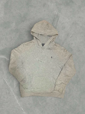Grå Ralph Lauren hoodie - Snygg grå hoodie från Polo Ralph Lauren. Klassisk design med huva, känguruficka och broderad logga på bröstet. Det är xl i barn storlek men sitter som en M eller en större S i herrstorlek. Den är i mycket bra skick och har bara används ett fåtal gånger. Vid frågor eller funderingar tveka inte att höra av er. 