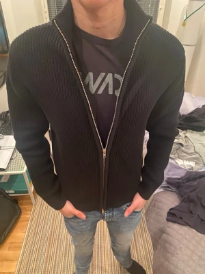 Svart stickad kofta med dragkedja - Säljer en svart stickad kofta med grov ribbad struktur och hel dragkedja framtill. Koftan har hög krage och långa ärmar, perfekt för lager-på-lager under kyliga dagar. Snygg och enkel att matcha med jeans eller t-shirt. Ifrån lager157