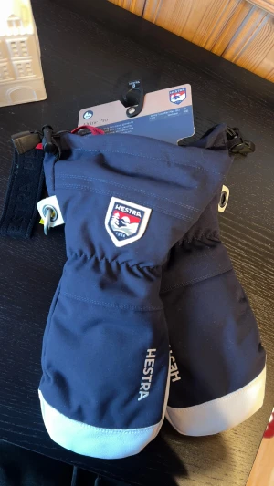 Hestra Alpine Pro skidhandskar - Säljer ett par helt oanvända mörkblå Hestra Alpine Pro skidhandskar. Handskarna har justerbar rem, metallring och är tillverkade i polyamid och skinn. Perfekt för vintern och skidåkning, med medföljande läderbalsam för vård.