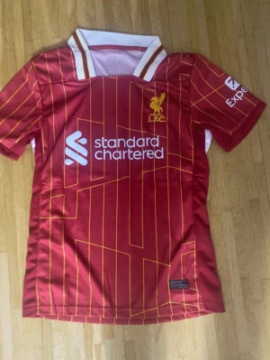 Röd Liverpool FC matchtröja - Säljer en röd Liverpool FC matchtröja med tunna gula ränder och vit krage. Tröjan har klubbens emblem på bröstet och sponsortryck i vitt. Tillverkad i polyester, perfekt för fotbollsfans som vill visa sitt stöd för laget.