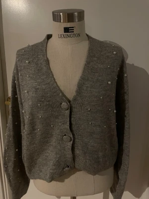 Grå kofta med pärlor H&M - Säljer en grå stickad kofta från Lexington med dekorativa pärlor över hela plagget. Koftan har v-ringad hals, stora knappar framtill och långa ärmar. Perfekt för dig som gillar detaljer och vill ha en mysig men snygg look.