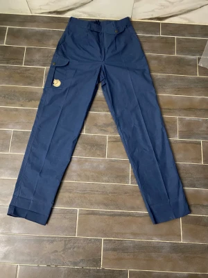 Blå cargopants från Fjällräven - Säljer ett par blå cargopants från Fjällräven med klassisk logga på benet. Byxorna har fickor med knappstängning på sidan och är tillverkade i slitstark bomull. Perfekt för dig som gillar outdoor och vill ha en clean look. Dom är som nya och har inga fläckar aldrig använt dom.