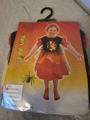 Fire Girl väst med eldtryck - Cool väst för barn med eldtryck på bröstet och puffiga röda ärmar. Västen har detaljer i rött, gult och svart som ger en eldliknande effekt. Perfekt för maskerad eller temafest. Materialet är 100% polyester och designen är färgstark och lekfull.