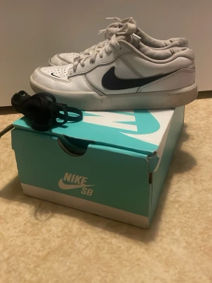 Nike SB vita sneakers med svart swoosh - Nike SB sneakers i vitt med svart swoosh på sidan. Skorna har snörning, perforerad tå och platt sula. Tillverkade i skinn och har en klassisk rund tå. Kommer med originalkartong och extra svarta skosnören. Perfekt för dig som gillar streetwear och skate. Använda ganska mycket finns skador på skorna men de går att använda, har försökt tvätta så mycket jag har haft tid med. Snöret som är på är ganska smutsigt, så vill du ha ett svart extra helt nytt snöre med skriv det till mig då! Ny pris 999kr!!