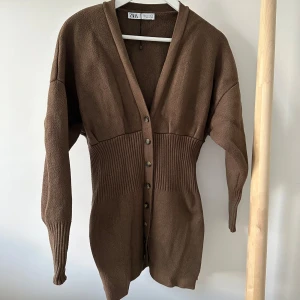Brun kofta från Zara med knappar - Säljer en brun lång kofta från Zara med djup v-ringning och knappar hela vägen framtill. Koftan är stickad med ribbade partier i midjan och vid ärmsluten, vilket ger en snygg och figurnära passform. 