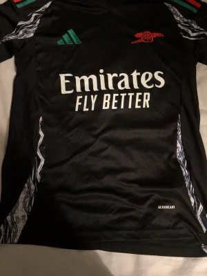 Arsenal bortatröja Adidas S - Säljer en svart Arsenal bortatröja från Adidas i storlek S, slim fit. Tröjan har gröna och röda detaljer vid kragen, vita mönster längs sidorna och tryck med 'Emirates Fly Better' på bröstet. Nummer 4 och namnet White på ryggen. Perfekt för fotbollsfans!