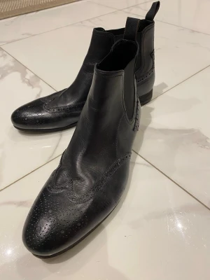 Svarta chelsea boots från Boss Orange - Snygga svarta chelsea boots från Boss Orange i äkta skinn med klassiska broguemönster och rund tå. Elastiska paneler på sidorna och dragflik bak gör dem enkla att ta på. Perfekt för dig som gillar stilrena och tidlösa skor med lite edge. 9/10 skick✅, riktigt stiliga  och är knappast använda.