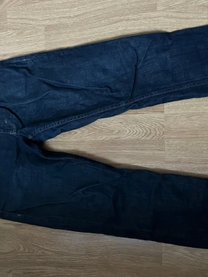 Mörkblå jeans från Bläck, storlek 31 - Säljer ett par mörkblå jeans från Bläck i storlek 31. Klassisk femficksmodell med raka ben och snygga kontrastsömmar. Jeansen har en stilren läderpatch med Bläck-logga bak i midjan. Materialet är slitstarkt denim i bomull.