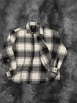 Rutig overshirt i svart och grått - Säljer en snygg rutig overshirt i svart, grått och vitt. Jackan har klassisk krage, knappar framtill och långa ärmar med knapp vid mudden. Perfekt att slänga över en hoodie eller t-shirt för en chill look.