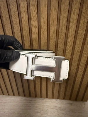 Vit skinnbälte med H-spänne - Snyggt vitt bälte i skinn med ett stort, silverfärgat H-spänne. Bältet har svart insida och tydliga sömmar längs kanterna. Perfekt för att lyfta din outfit med en lyxig detalj.