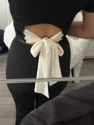 Svart croppad topp med rosett - Säljer en svart croppad topp med öppen rygg och stor beige rosett som knyts baktill. Har aldrig använt denna toppen/ ny. Säljs på grund av flytt