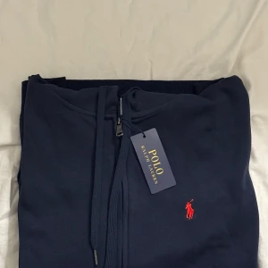 Marinblå hoodie från Polo Ralph Lauren - Snygg marinblå hoodie från Polo Ralph Lauren med dragkedja framtill och klassisk huva. Tröjan har snörning vid huvan och den ikoniska röda Polo-loggan broderad på bröstet. Perfekt för en chill och stilren look.