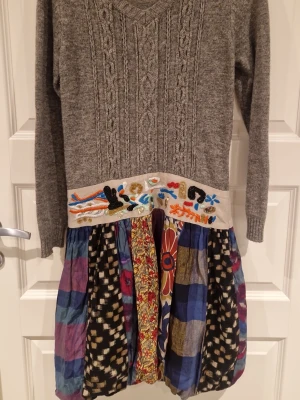 Desigual färgglad mönstrad klänning - Unik klänning från Desigual med grå stickad överdel, v-ringning och långärmad. Midjan har broderade detaljer och kjolen är färgglad med blommigt, rutigt och prickigt mönster. Materialmix av linne, bomull, nylon, ull och polyester. Perfekt för dig som gillar kreativa och lekfulla plagg.