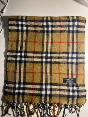 Rutig Burberry halsduk  i kashmir - Säljer en klassisk rutig väst från Burberry i 100% kashmir. Färgerna är beige, svart, vit och röd med det ikoniska rutmönstret. Västen har fransar längst ner och är tillverkad i England. Perfekt för dig som gillar lyxiga och tidlösa plagg.