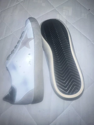 Golden Goose Superstar sneakers silver - Golden Goose Superstar sneakers i vitt med silverfärgade detaljer på hälen och grå stjärna på sidan. Skorna har grå snörning och en platt sula med mönstrad undersida. Märket syns tydligt på insidan och sidan av skon. Perfekt för dig som gillar en clean och cool stil. Både män och kvinnor kan använda 