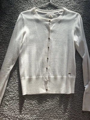 Vit kofta från Holly & Whyte - Snygg vit kofta från Holly & Whyte by Lindex i storlek S. Koftan har rund hals, långa ärmar och  guldiga knappar framtill. Materialet är mjukt och tunt, perfekt för lager-på lager. Funkar alla årstider och är en fin casual tröja som ändå blir lite extra. Använd några gånger men säljer pga att den blivit för liten.❤️❤️