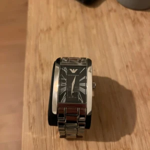 Armani klocka  - Bilderna visar en Emporio Armani klocka med svart urtavla och silverfärgad länkarmband, samt tillhörande förvaringsask som har defekt. 