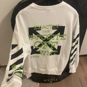 Vit sweatshirt från Off-White med tryck - Vit sweatshirt från Off-White med grönt och svart grafiskt tryck på både fram- och baksida samt ärmar. Tröjan har rund hals, lång ärm och coola diagonala ränder med bokstäver och siffror. Materialet är mjuk bomull, perfekt för streetwear-stil.