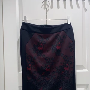 Mörkröd kjol med spetsdetaljer svart och råd - Snygg mörkröd kjol med svarta spetsdetaljer och broderat blommönster. Kjolen har en tight passform och är knälång med dekorativ kant nertill. Materialet känns lyxigt och har en elegant look med dragkedja bak. Perfekt för dig som gillar unika detaljer och stil.