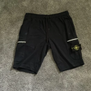 Svarta shorts från Stone Island - Säljer ett par svarta shorts från Stone Island med dragsko i midjan och stora sidofickor. På ena benet sitter den klassiska Stone Island-loggan. Shortsen har vita detaljer vid fickorna och är tillverkade i mjukt material, perfekta för träning eller chill.