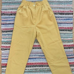 Gula högmidjade kostymbyxor  - Gula kostymbyxor med hög midja och klassisk rak passform. Byxorna har hällor för bälte, med veck framtill. Perfekta för dig som vill sticka ut med färg och ändå hålla det stilrent. Storlek 34-36. 