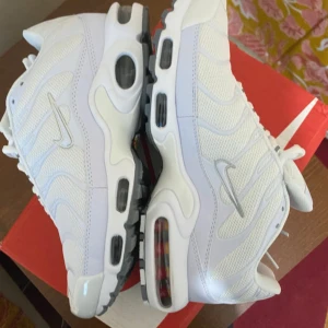 Nike Air Max Plus vita sneakers - Nike Air Max Plus i helvitt med mesh och syntetpaneler, ikoniska vågformade detaljer och synliga Air Max-enheter i sulan. Snygg design med grå och svarta detaljer på yttersulan och klassisk Nike-logga på sidan. Perfekta för dig som gillar streetstyle och komfort.