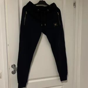 Svarta mjukisbyxor MR Originals S - Svarta mjukisbyxor från MR Originals med guldfärgade dragkedjor på fickorna och snörning i midjan. Byxorna har en liten guldfärgad logga framtill och ribbade muddar vid bensluten. Perfekta för en chill och avslappnad stil.