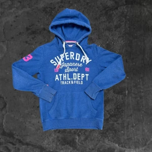Blå hoodie från Superdry med tryck - Säljer en blå hoodie från Superdry med vit och rosa text på bröstet och siffror på ärmarna. Tröjan har huva med snörning, känguruficka och ribbade muddar. Perfekt för en sportig och avslappnad stil.