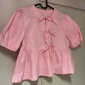 Rutig rosa blus med volang och knyt - Supersöt blus i ljusrosa och vitt med rutigt mönster. Blusen har puffiga korta ärmar, volang nedtill och tre knytband framtill. Materialet känns som bomull och ger en luftig känsla. Perfekt för dig som gillar gulliga detaljer och en feminin stil.