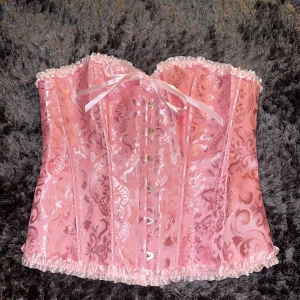 Rosa korsett med snörning och rosett - Säljer en glansig rosa korsett med vackert broderat mönster och vita volangdetaljer längs kanterna. Framtill finns en söt rosett och knappar, medan baksidan har snörning för perfekt passform. Materialet har en satinlik finish som ger en lyxig känsla. Helt ny endast testad förpackningen finns kvar. Den var för stor för mig och hann inte returnera den i tid. 