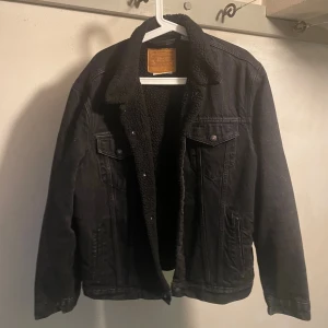 Svart jeansjacka med teddyfoder Levi's XL - Svart jeansjacka från Levi's Premium i storlek XL med mjukt svart teddyfoder på insidan och kragen. Klassisk modell med två bröstfickor med knappar och sidofickor. Perfekt för kyliga dagar och har en cool, tidlös look.
