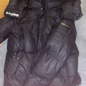 Svart pufferjacka från Gabos London - Säljer en svart pufferjacka från Gabos Jeans med hög krage och dragkedja framtill. Jackan har stora fickor med dragkedja och tydliga logodetaljer på ärmen och bröstet. Perfekt för kalla dagar när du vill hålla stilen. Har ett litet hål i armhålan men annars fint skick