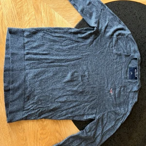 Mörkgrå stickad tröja från Hollister - Snygg mörkgrå stickad tröja från Hollister med diskret logga broderad på bröstet. Tröjan har rund hals, ribbade muddar och långärmad passform. Perfekt för dig som gillar enkel och stilren look. Passar dig som vill ha en normal passning.