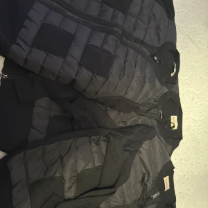 Moncler Cardigans Lisa  - Säljer en svart pufferjacka från Moncler med quiltad design och ribbade detaljer. Jackan har hel dragkedja framtill, två fickor och klassisk Moncler-logga på etiketten. Perfekt för dig som vill ha en stilren och sportig look.