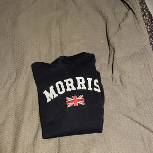 Mörkblå hoodie från Morris - Säljer en svart hoodie från Morris med en stor logotyp och en brittisk flagga på framsidan. Tröjan har en klassisk huva med dragsko och långa ärmar. Perfekt för en avslappnad stil.