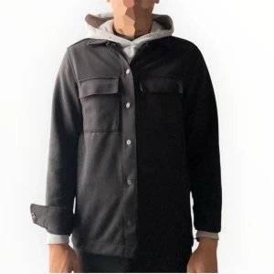 Overshirt - MQ - Stilren svart overshirt med klassisk krage, två stora bröstfickor med lock och tryckknappar framtill. Jackan har raka ärmar med knappdetalj vid ärmslut och är tillverkad i ett mjukt material som ger en clean look. (Modellen är 178 och väger 72 kg). Priset är inte hugget i sten