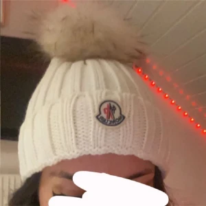 Vit Moncler mössa med pälsboll - Vit ribbstickad mössa från Moncler med stor fluffig pälsboll på toppen. Mössan har Moncler-logga framtill och är tillverkad i ett mjukt, stickat material. Perfekt accessoar för kalla dagar och ger en lyxig vinterlook.